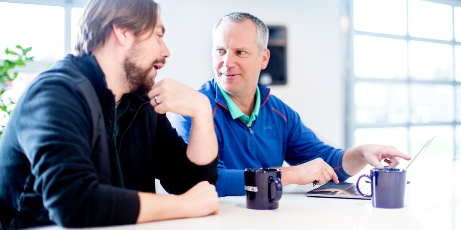 two men discussing at a table with laptop and coffee mugs