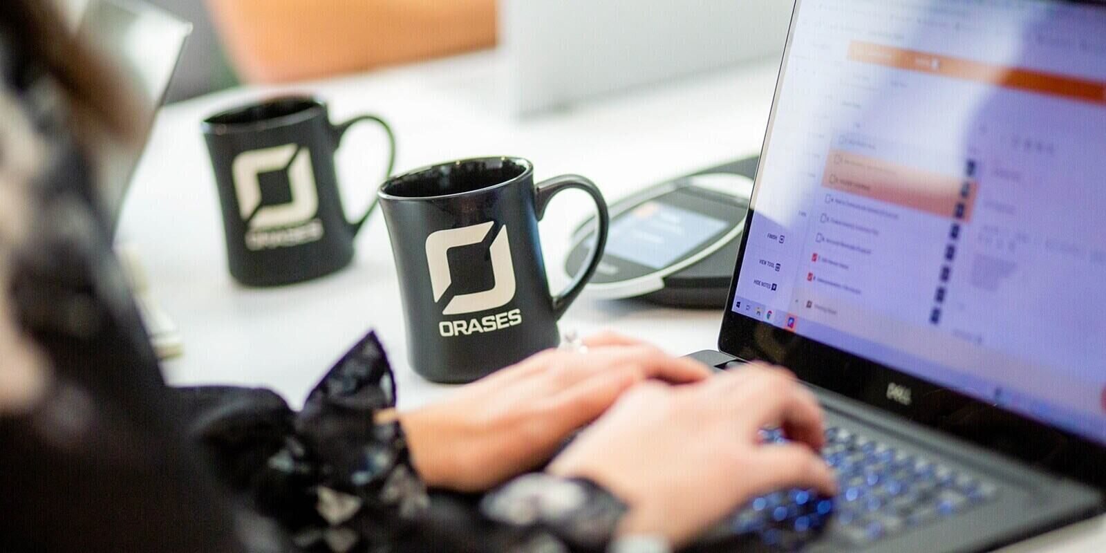 person typing on a laptop keyboard with two branded Orases coffee mugs and a conference call speaker in the background